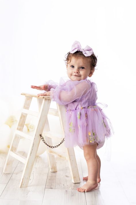 Una niña con un vestido y un lazo de color lila está de pie junto a una escalera blanca sobre un fondo de estudio luminoso.