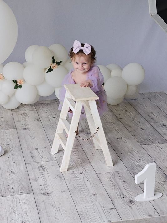 Niña con un traje y un lazo color lavanda posando en una escalera blanca, con globos blancos y un gran “1” al fondo.