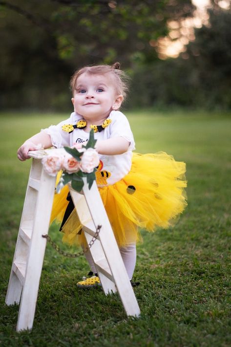 Bebé sonriente con un tutú amarillo apoyado en una escalera blanca en un jardín cubierto de hierba.