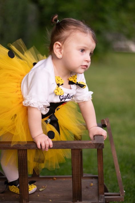 Una niña pequeña con un tutú amarillo está apoyada en un banco de madera al aire libre, mirando hacia un lado.