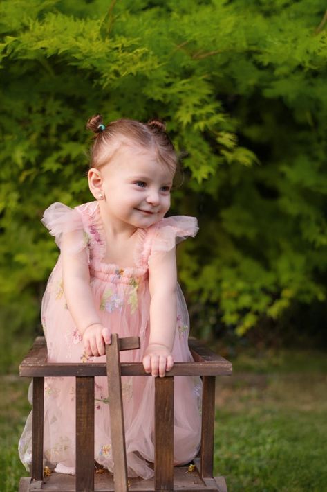 Niña sonriente con un vestido rosa apoyada en una valla de madera al aire libre, con vegetación verde de fondo.