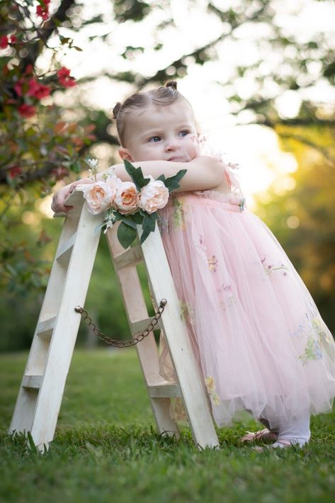 Niña con un vestido rosa apoyada en una escalera decorada con flores al aire libre sobre el césped.