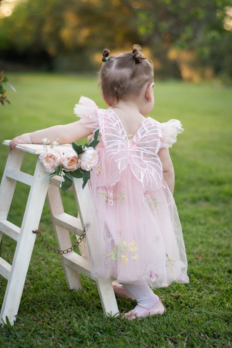 Niña con un vestido rosa apoyada en una silla blanca en un jardín cubierto de hierba.