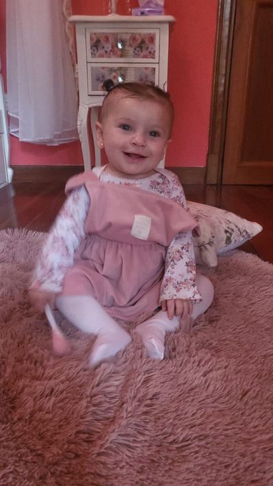 Bebé sonriente con un conjunto rosa sentado sobre una alfombra rosa mullida en una habitación rosa.