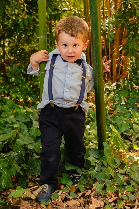 Un niño, de pie en un bosquecillo de bambú, viste una camisa clara y tirantes oscuros, y sostiene un tallo de bambú.