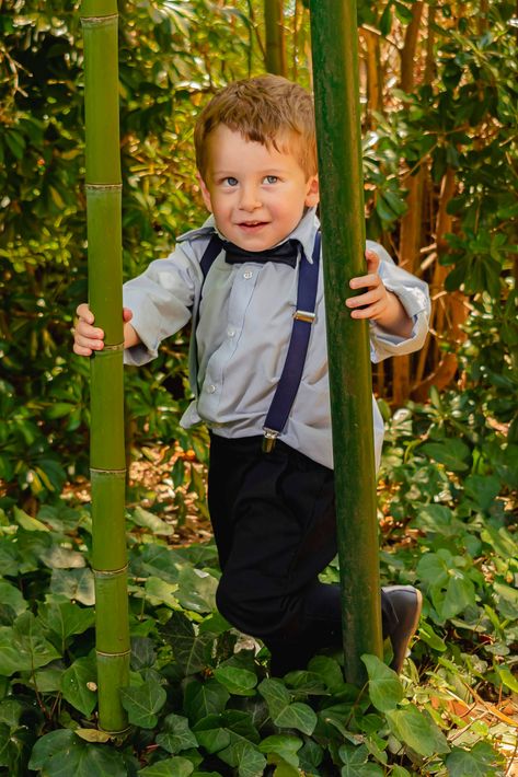 Un niño sonríe en un columpio de bambú en un jardín exuberante, vistiendo una camisa gris y un mono oscuro.