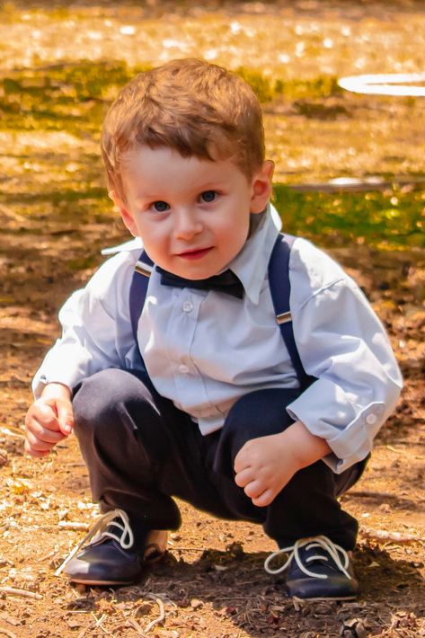 Un niño pequeño, agachado al aire libre en un camino de tierra, viste una camisa blanca, pajarita, tirantes 