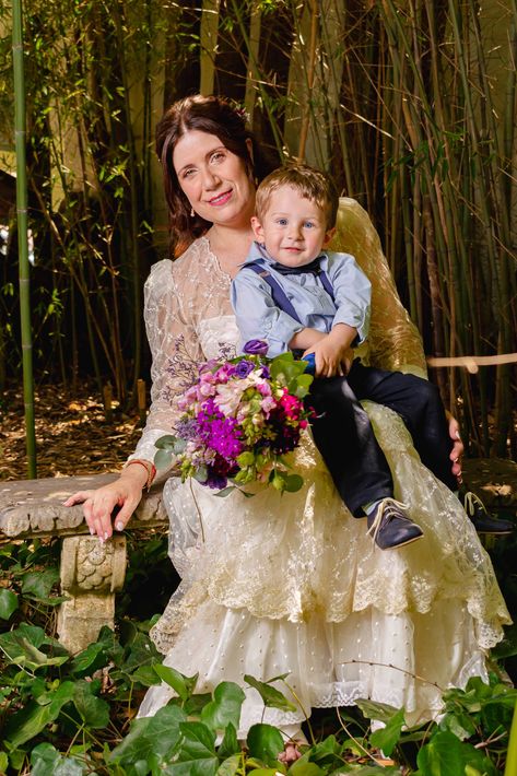 Mujer con vestido color crema, sosteniendo a un niño y un ramo de flores moradas, sentada al aire libre entre bambú.