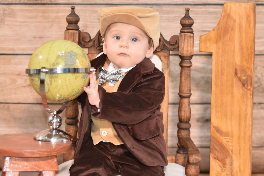 Un niño con un traje marrón y un sombrero color canela, sentado en un banco de madera junto a un globo terráqueo amarillo.