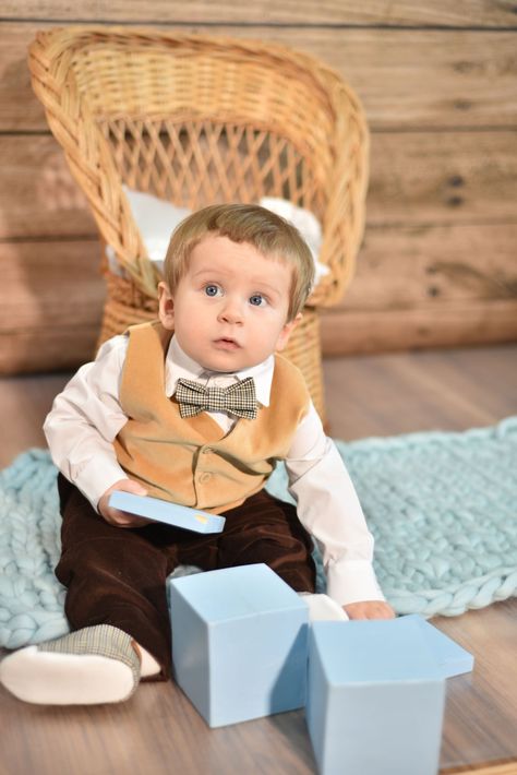 Un niño pequeño con pajarita está sentado en un suelo de madera con bloques azules y una silla de mimbre detrás.