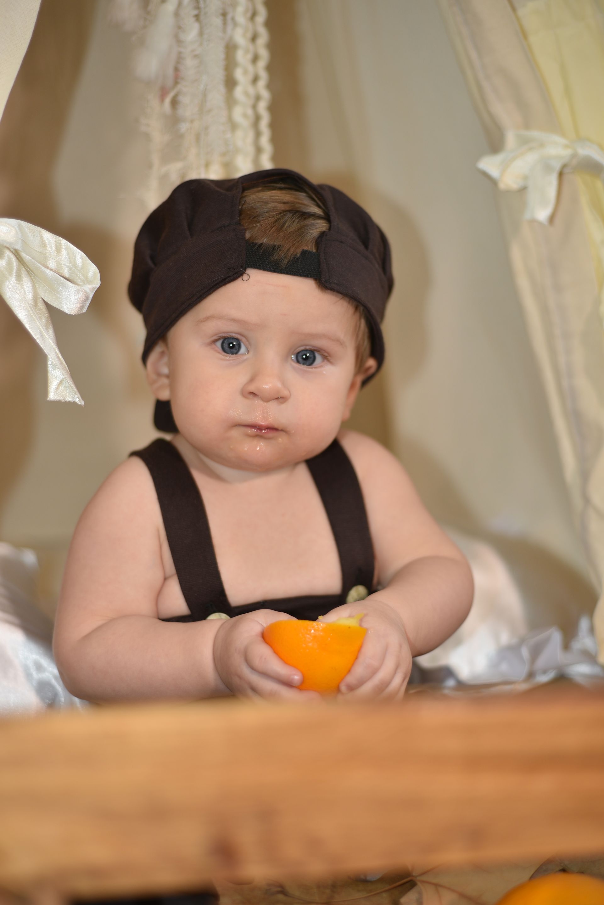 Un bebé con gorro oscuro y tirantes sostiene un objeto naranja en un estudio decorado con suaves cortinas.