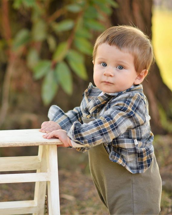 Un niño pequeño con camisa de cuadros y pantalones verde oliva está apoyado en un banco blanco al aire libre.