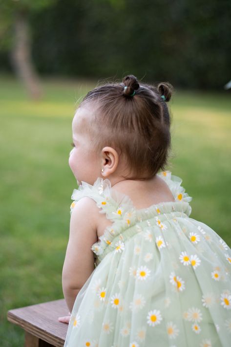 Niña con un vestido verde claro estampado de margaritas sentada al aire libre en un banco, mirando hacia otro lado.