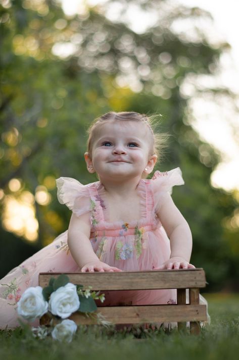 Bebé sonriente con un vestido rosa sentado en una caja de madera con flores blancas al aire libre.