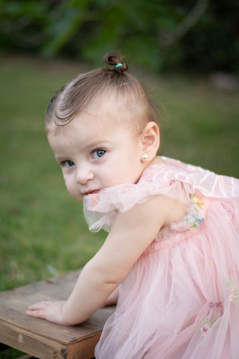 Una niña pequeña con un vestido rosa, apoyada en un banco de madera al aire libre, mira hacia la cámara.