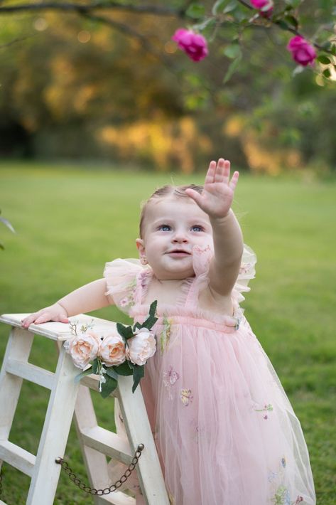 Una niña pequeña con un vestido rosa estira los brazos junto a un banco blanco en un jardín de flores.