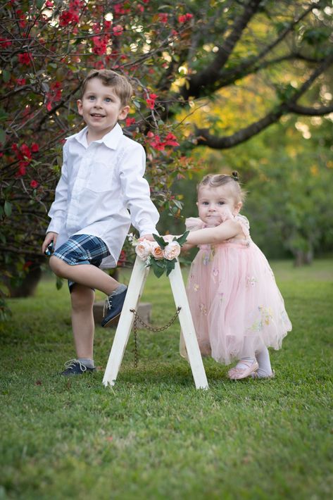 Dos niños con atuendos blancos y rosas posando sobre el césped junto a un árbol en flor al aire libre.