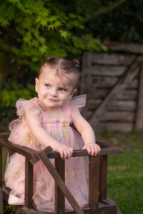 Una niña pequeña con un vestido rosa, apoyada en una silla de madera al aire libre, mira hacia un lado.