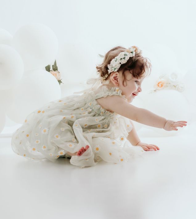 Niña con un vestido color crema gateando sobre el suelo blanco de un estudio, extendiendo el brazo hacia .