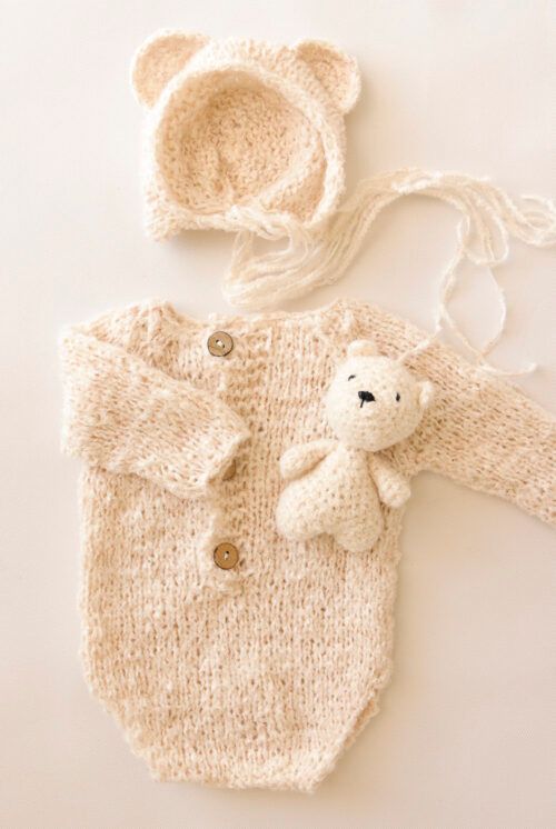 Conjunto de bebé de punto color crema con capucha de oso, gorro y un pequeño osito de peluche sobre un fondo claro.