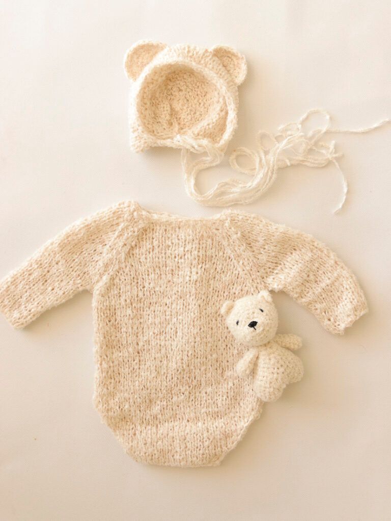 Conjunto de bebé de punto color crema con capucha de oso, gorro y un pequeño osito de peluche sobre un fondo claro.