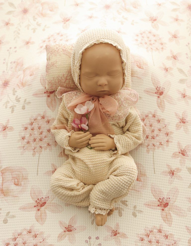 Conjunto de punto color crema para bebé con gorro rosa, zapatitos y flores sobre un fondo beige suave.