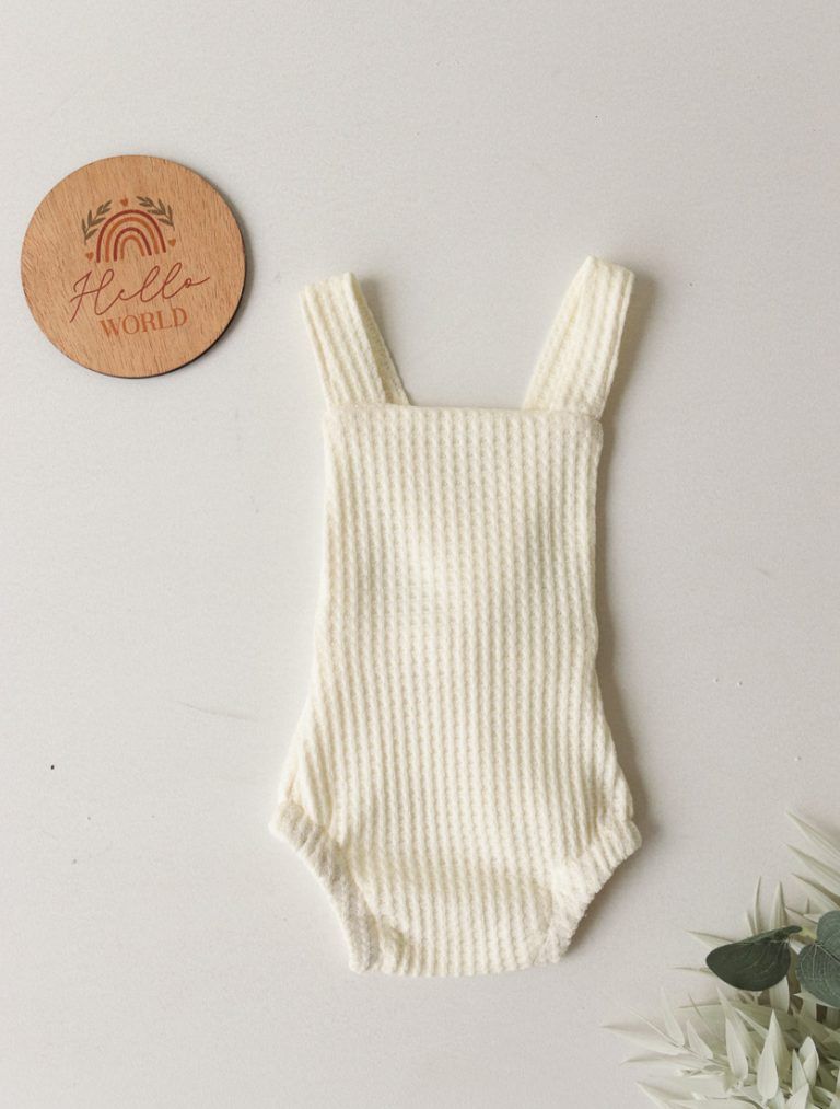 Pelele de bebé de punto acanalado color crema con tirantes, junto a una etiqueta redonda de madera sobre un fondo claro.