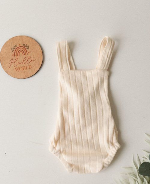 Pelele de bebé de punto acanalado color crema con tirantes, junto a una etiqueta redonda de madera sobre un fondo claro.