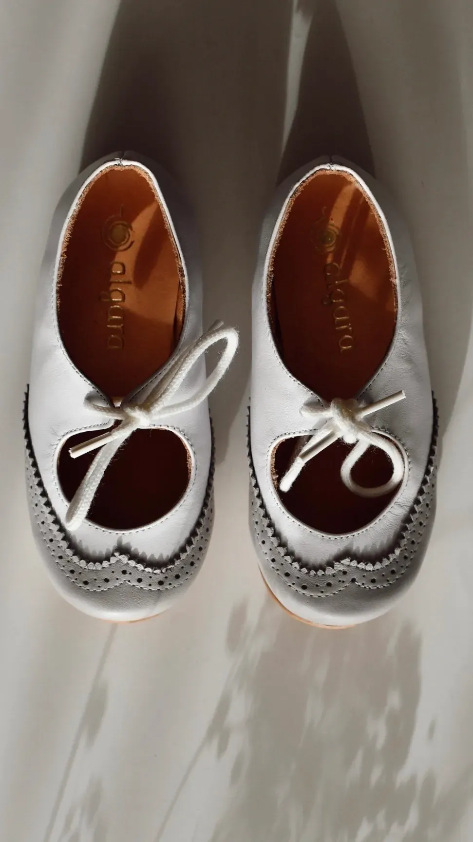 Un par de zapatos blancos con cordones y plantillas marrones, colocados uno al lado del otro sobre una superficie clara.