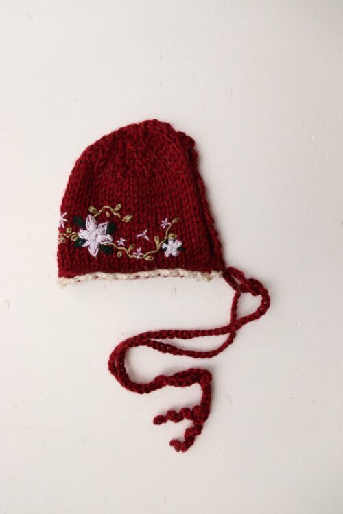 Gorro de bebé de punto rojo y blanco con estampado floral y cintas para atar al mentón sobre fondo blanco.