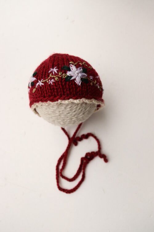 Gorro de bebé de punto rojo y blanco con estampado floral y cintas para atar al mentón sobre fondo blanco.