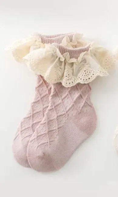 Calcetín rosa de bebé con puño de volantes de encaje color crema sobre fondo blanco.