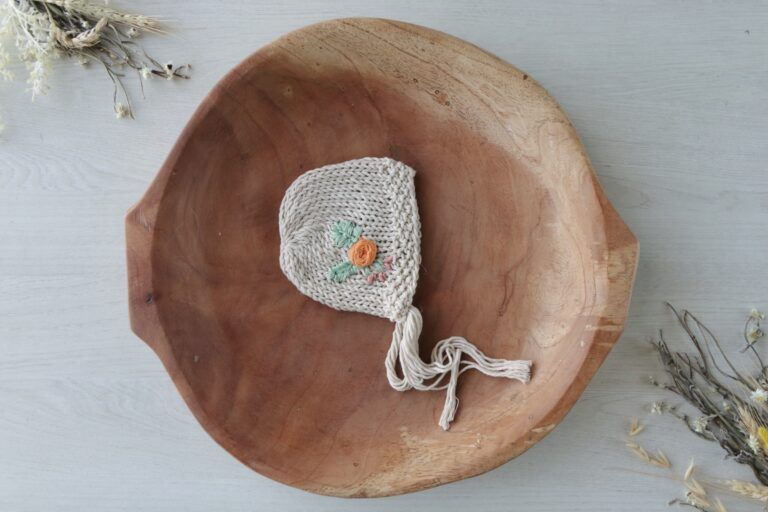 Gorro de bebé de punto color crema con cordones para atar sobre una cabeza de muñeca redonda tejida, sobre un fondo blanco.