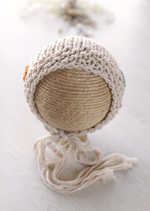 Gorro de bebé de punto color crema con cordones para atar sobre una cabeza de muñeca redonda tejida, sobre un fondo blanco.