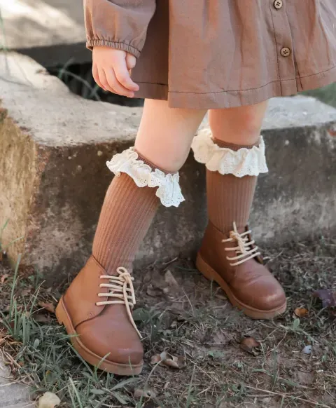 Niño con abrigo marrón y botas, de pie al aire libre sobre una cornisa de piedra.