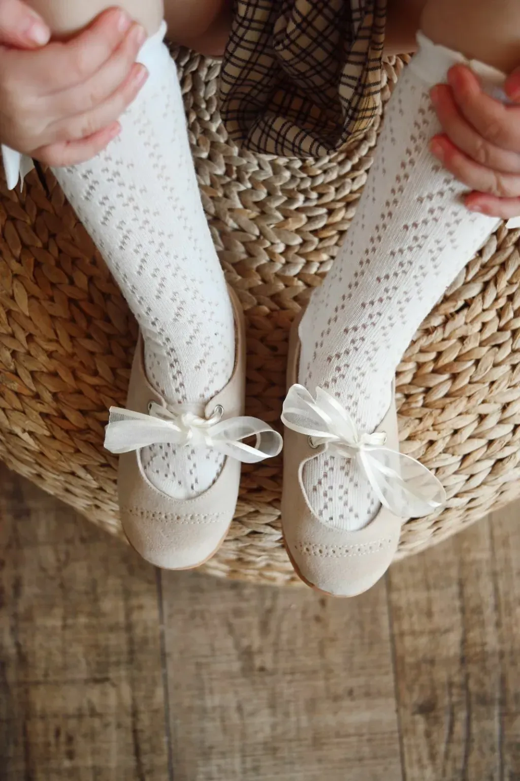 Patucos de bebé color crema con lazos blancos y calcetines de punto sobre una silla de mimbre, sobre un suelo de madera.