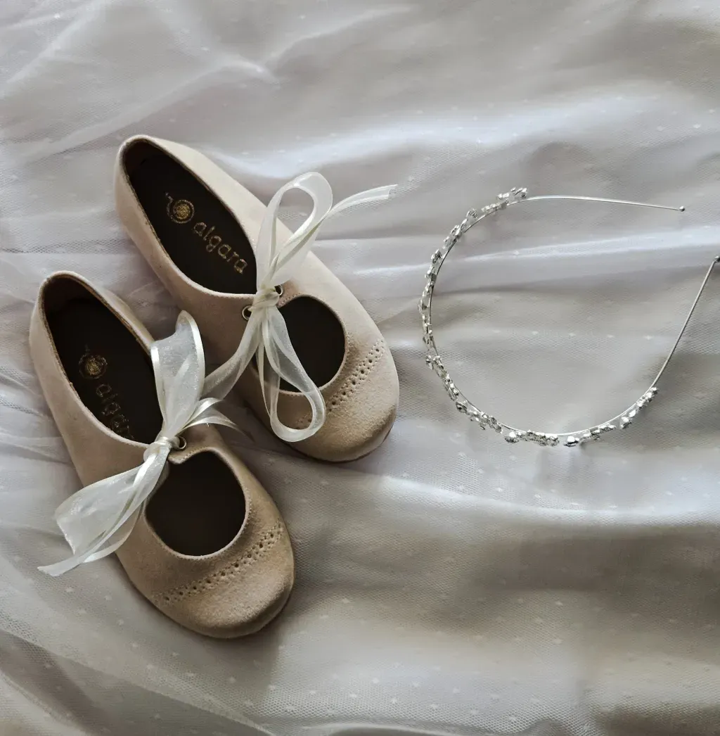 Un par de bailarinas beige con cordones y un delicado collar de plata sobre un fondo de tela blanca.