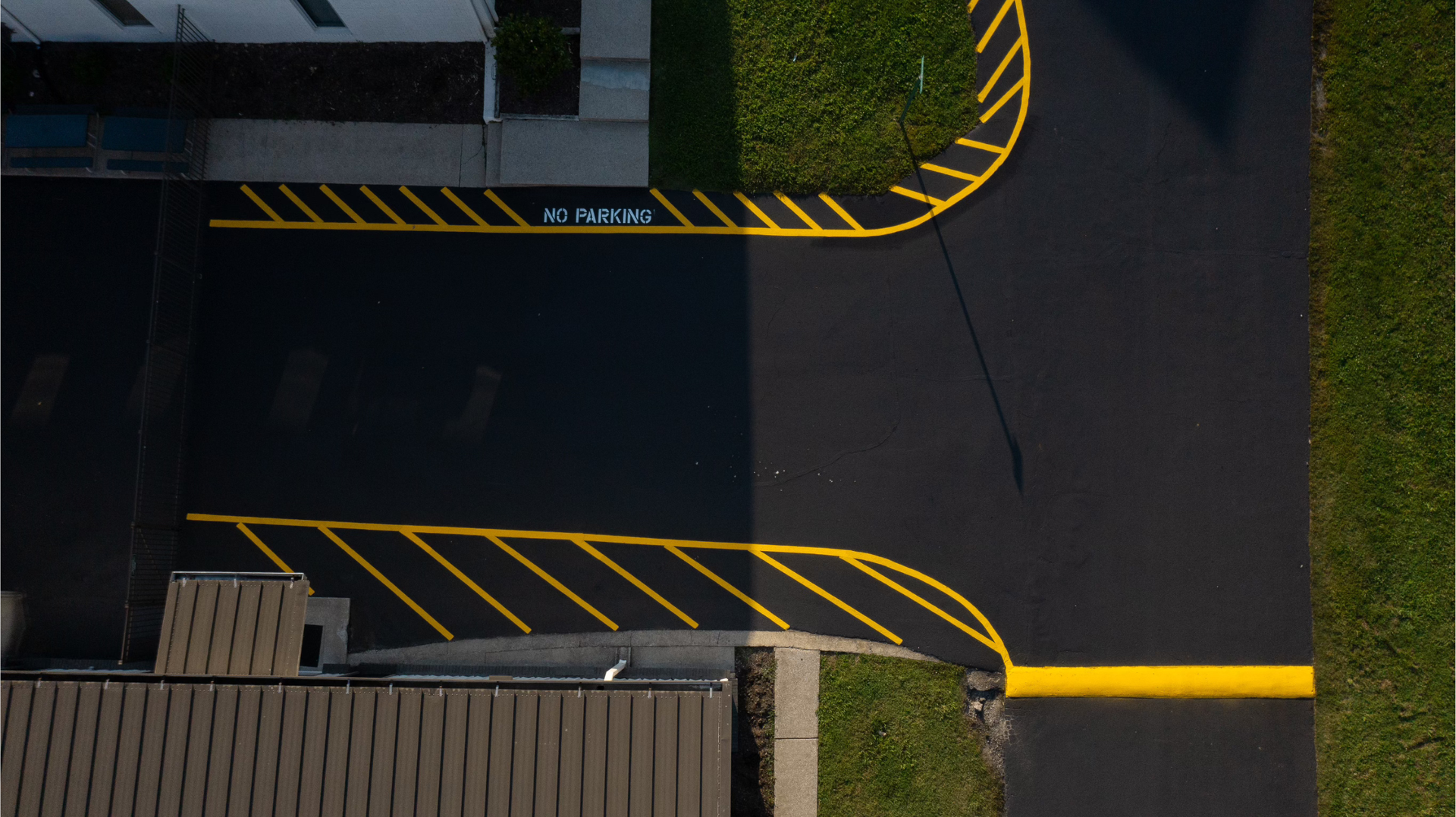 Overhead view of a paved area with yellow no parking markings and grass. Building edges visible.
