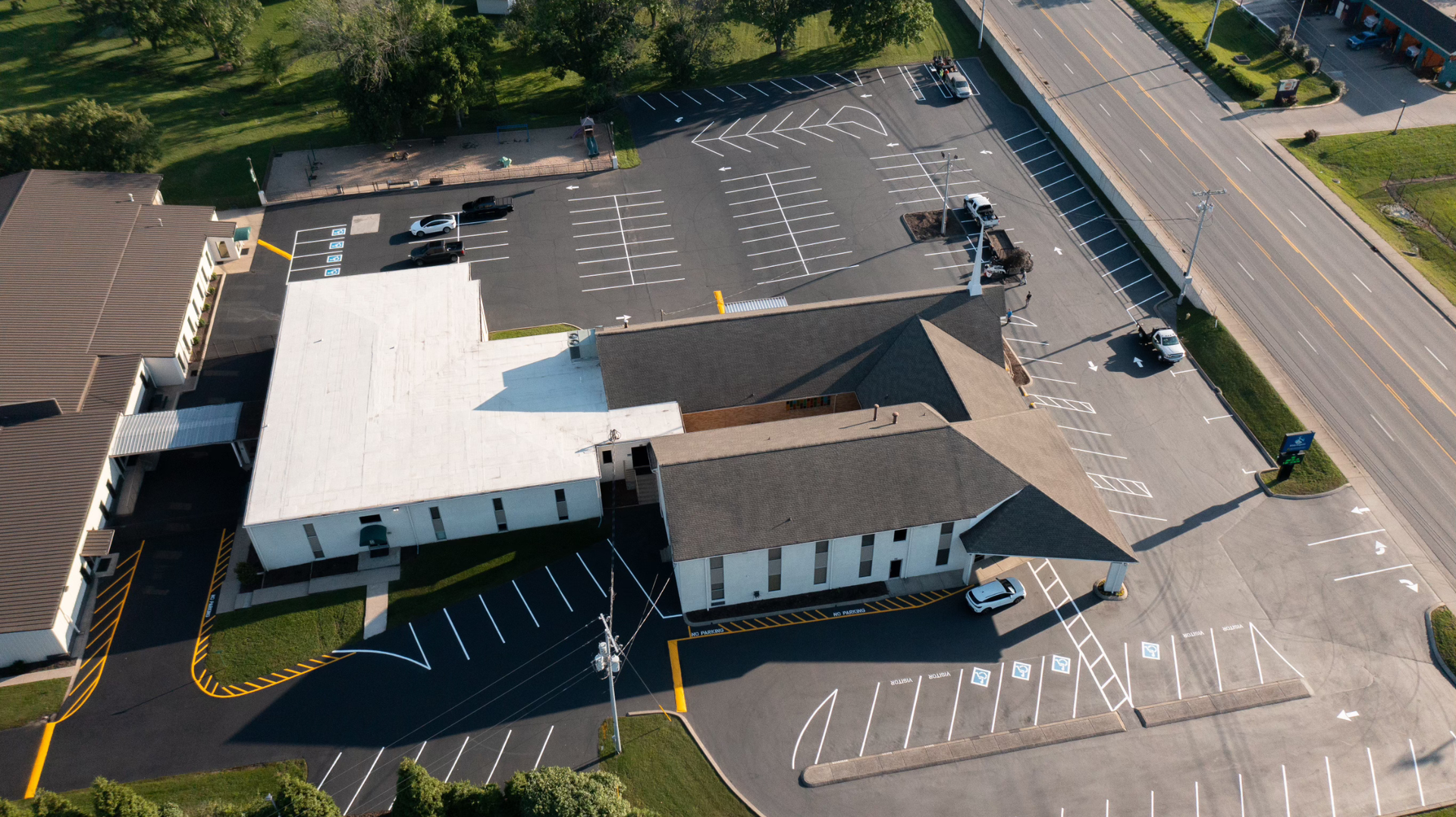 Aerial view of a church with a parking lot. White building with dark roof, surrounded by asphalt and green lawn.