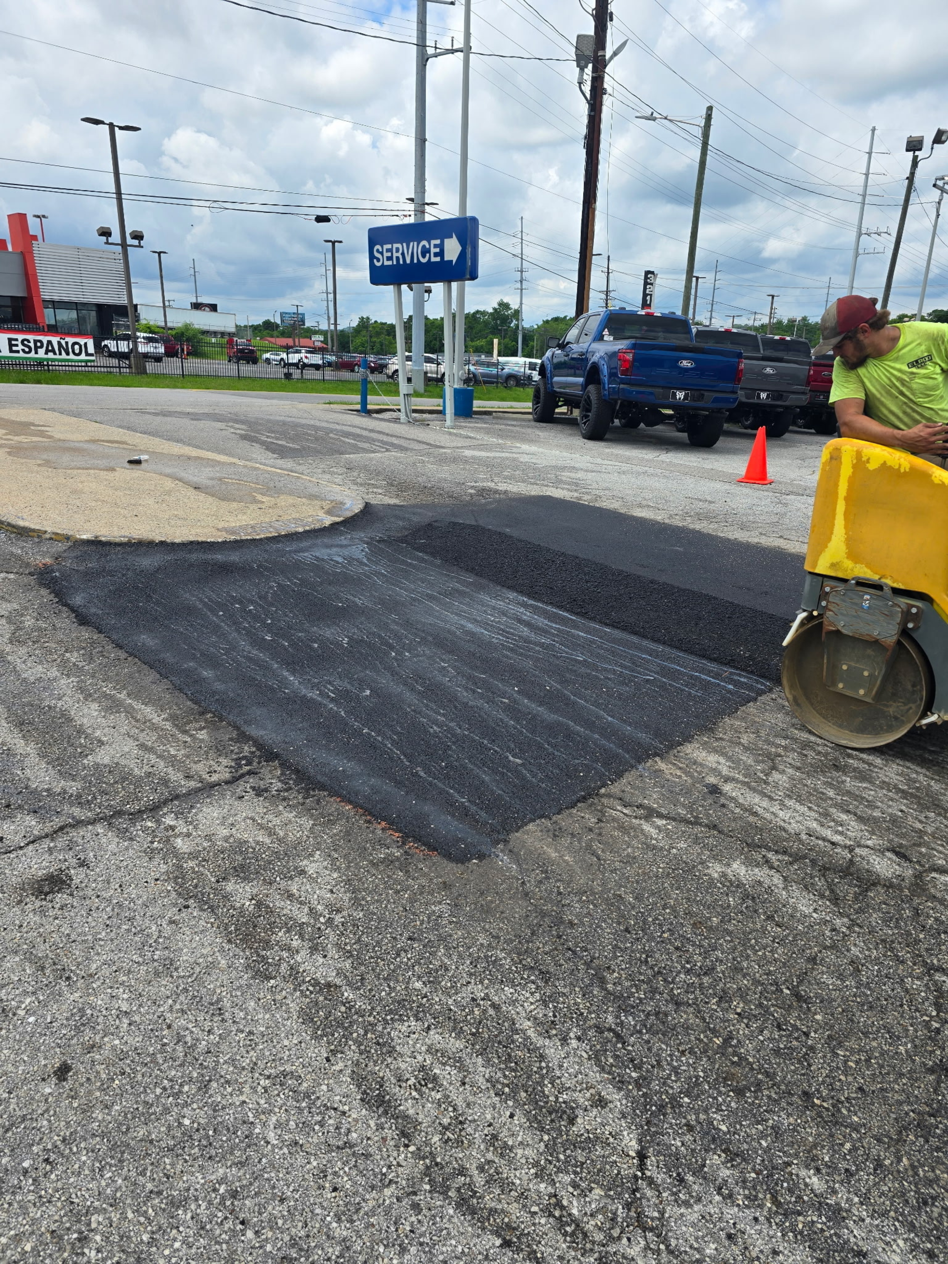 Asphalt patch being compacted by a small roller. Man in neon shirt watches. Outdoor setting.