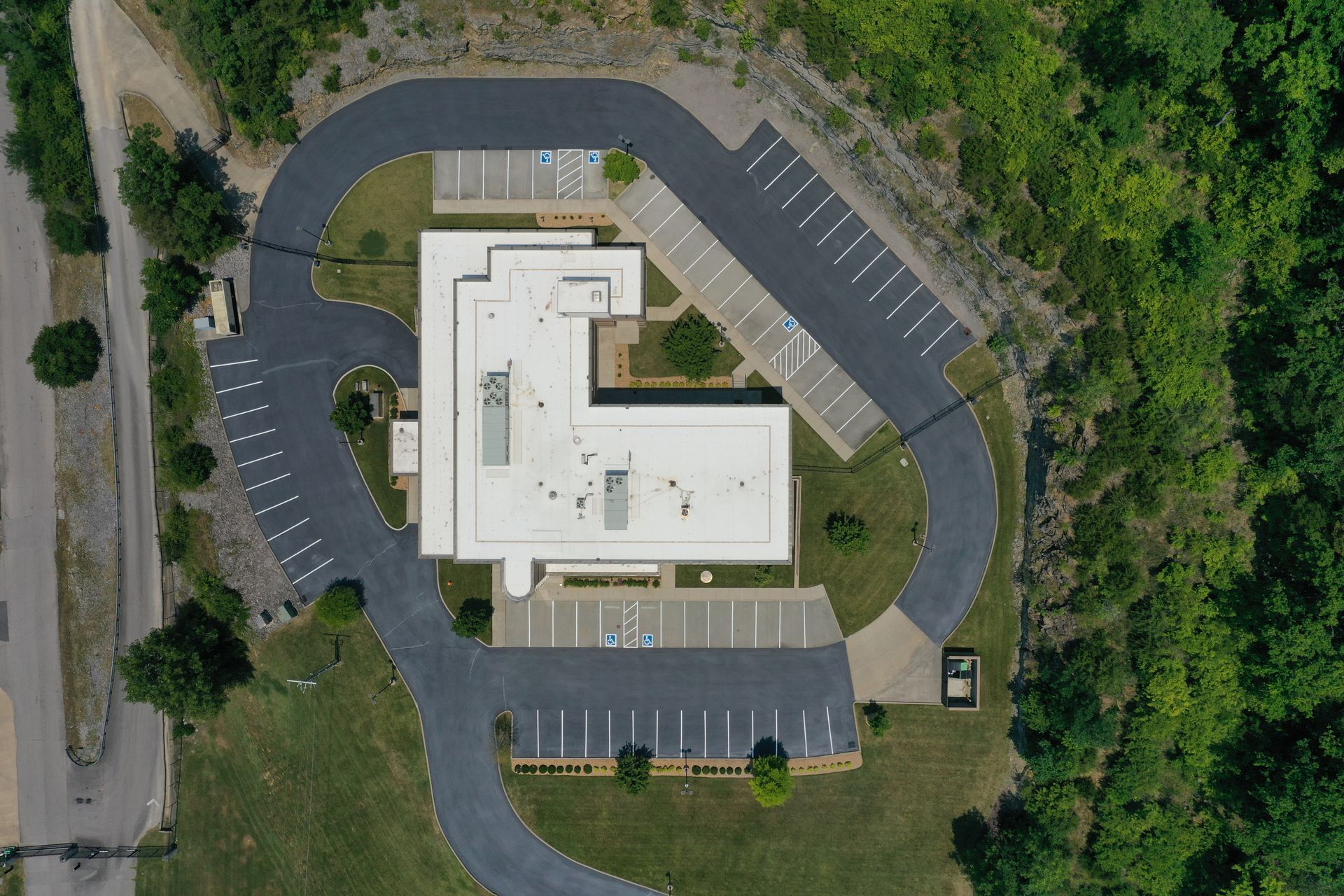 Aerial view of a white, L-shaped building with surrounding black asphalt parking and road, nestled in green trees and grass.