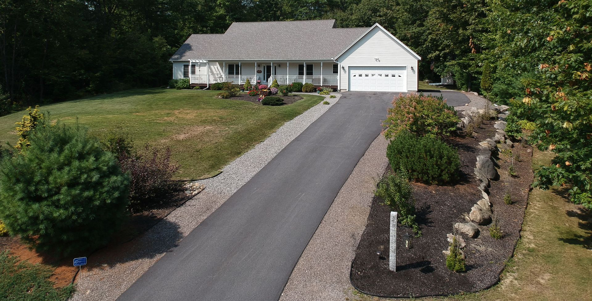 Ranch-style house with gray roof, white trim, and a long asphalt driveway bordered by landscaping.
