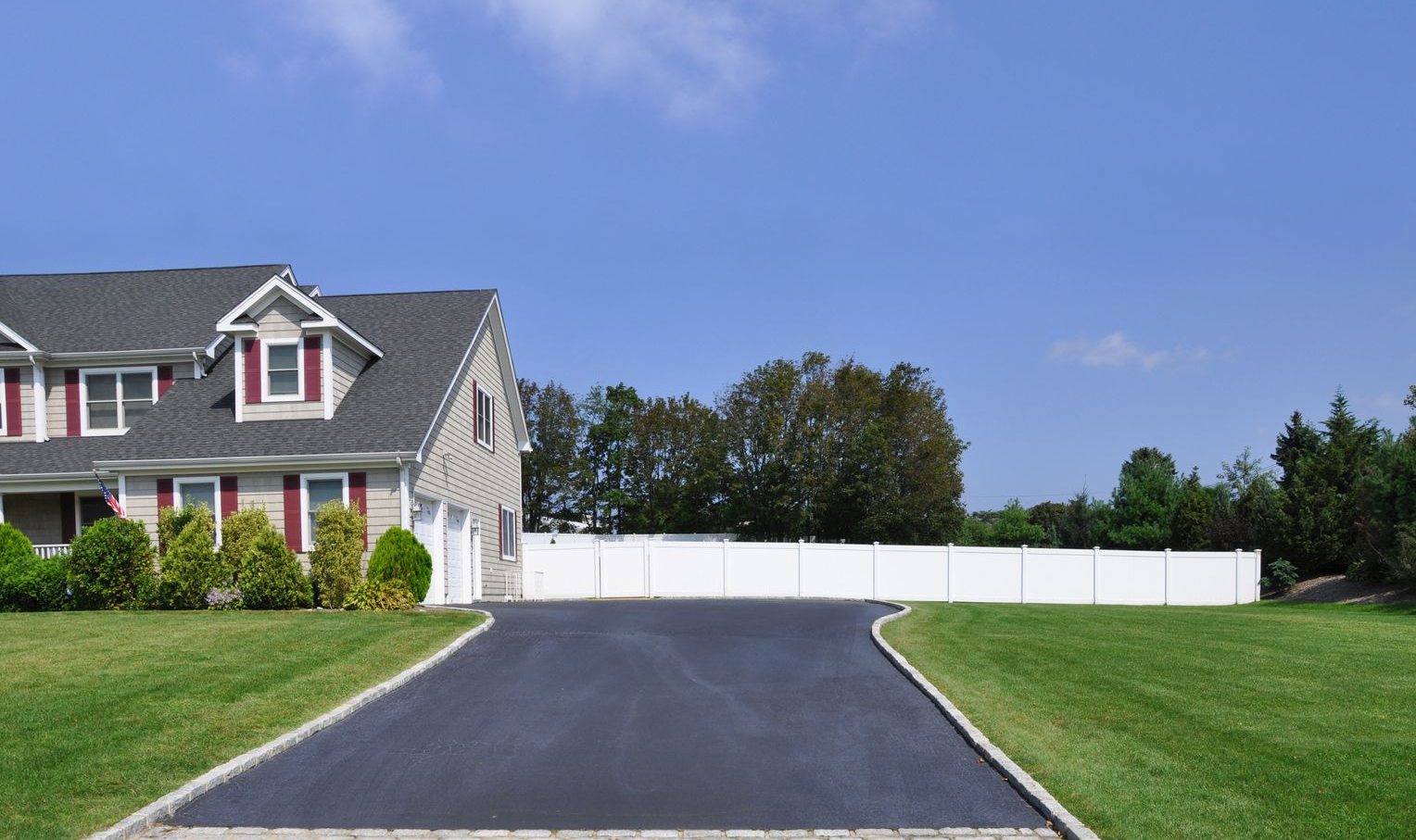 House with gray roof, black asphalt driveway, and white fence against a blue sky with clouds.