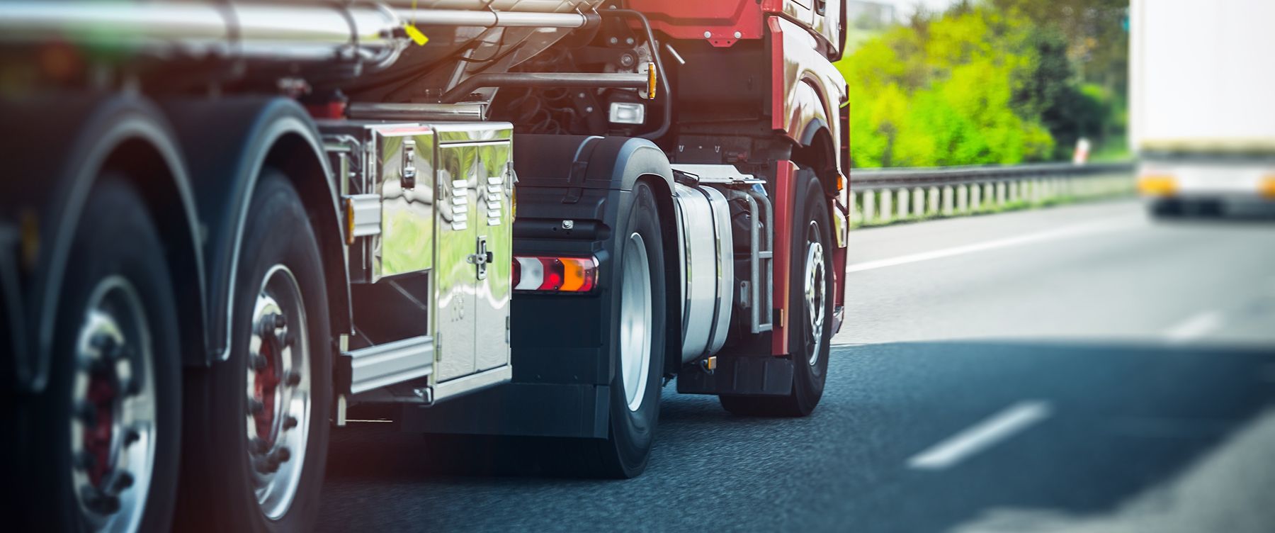Red semi-truck driving on a highway beside a guardrail, with a blurred car ahead.