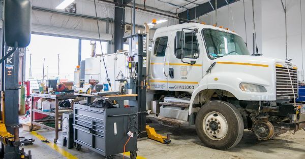 White truck in a repair shop, with open bay doors. Tools, equipment, and workbenches are visible.
