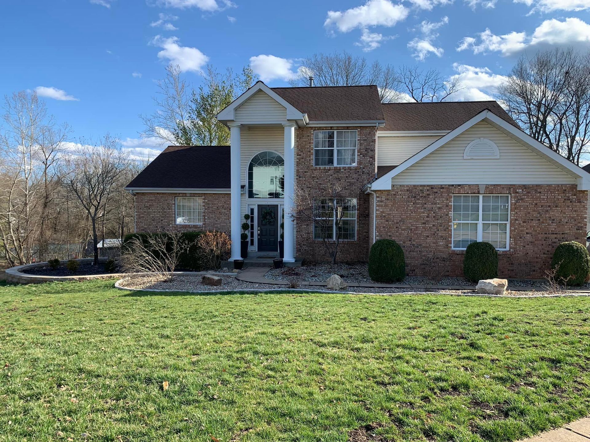 Two-story brick house with arched entrance, brown roof, and green lawn on a sunny day.