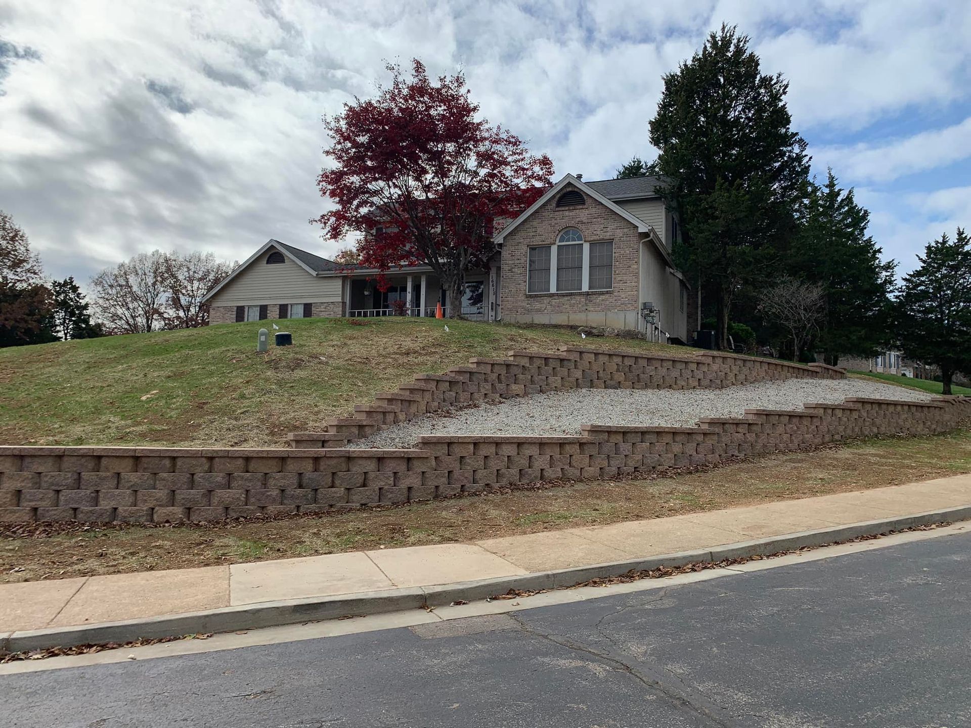 Brick retaining wall with gravel and grass leading to a two-story house, a tree with red leaves visible.