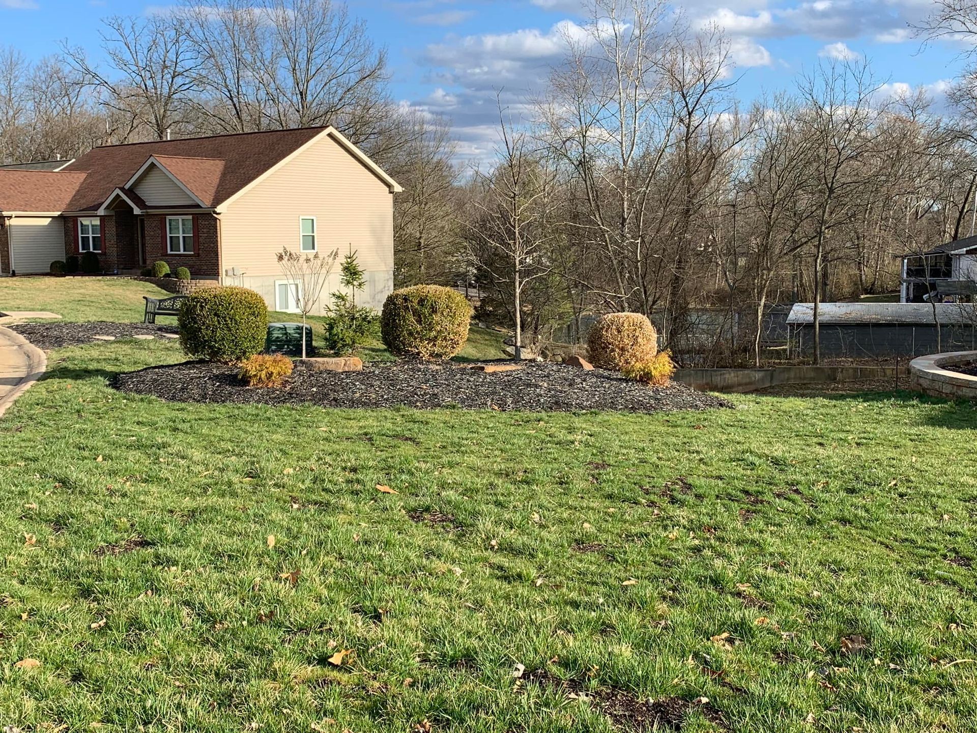 House with lawn, bushes, and trees in front. Cloudy blue sky.