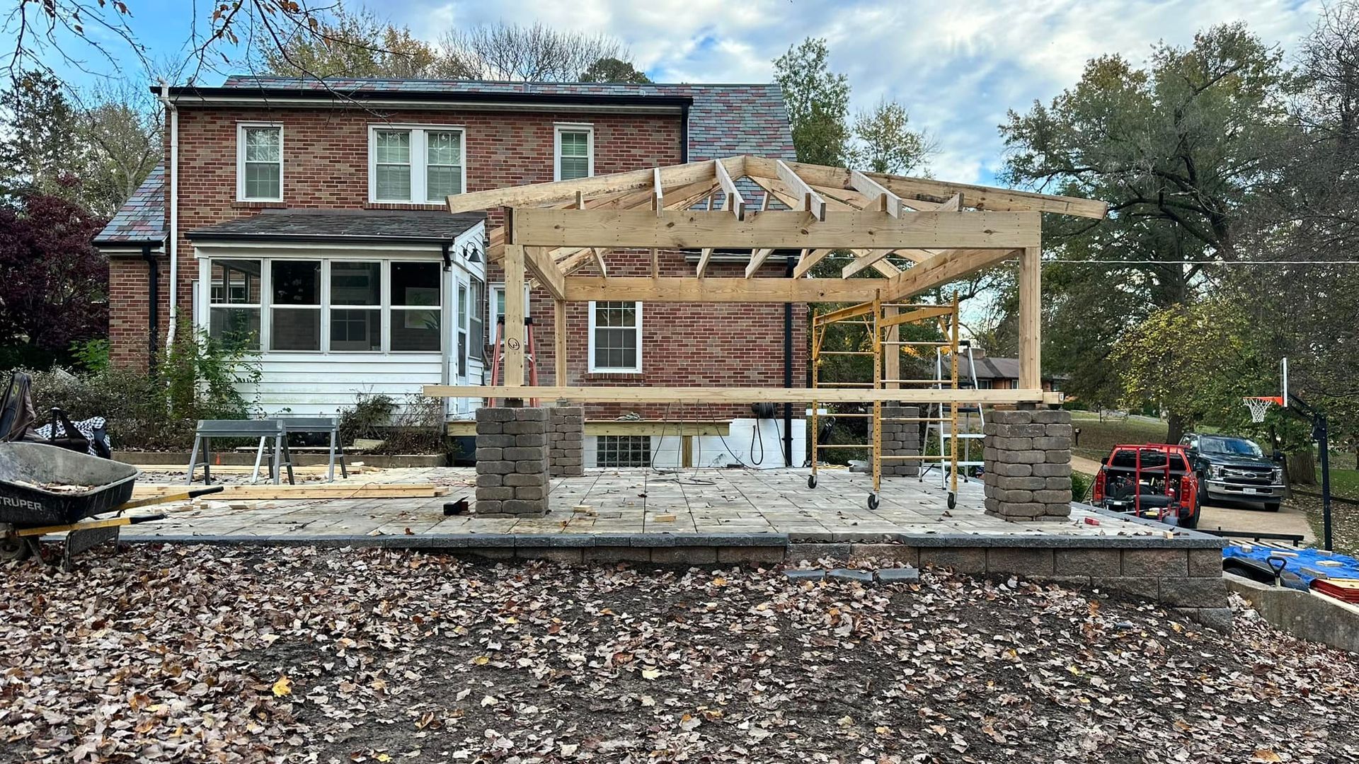 Construction of a wooden gazebo addition on a brick home with an unfinished patio and foliage.