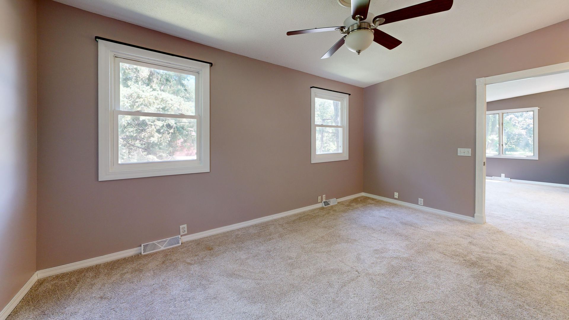 Empty beige carpeted bedroom with three windows, ceiling fan, and doorway to a bright adjoining room